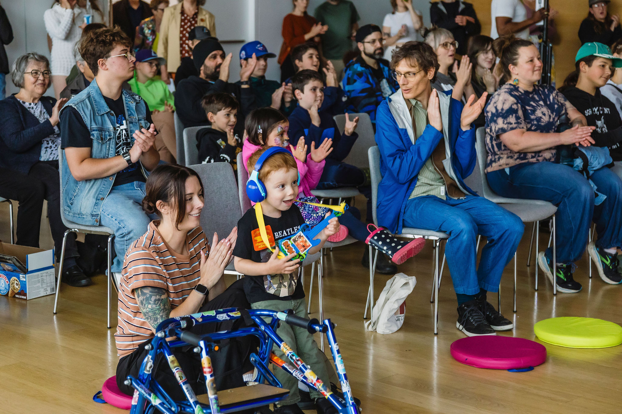 A seated audience of all ages applauds, with a child in blue ear defenders in the front row holding a toy instrument beside an adult.